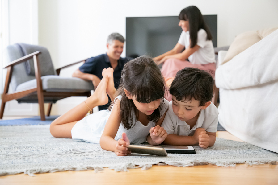 Cute little kids lying on floor in living room and using digital gadgets with learning apps while parents laughing in background. Internet communication or digital technology concept