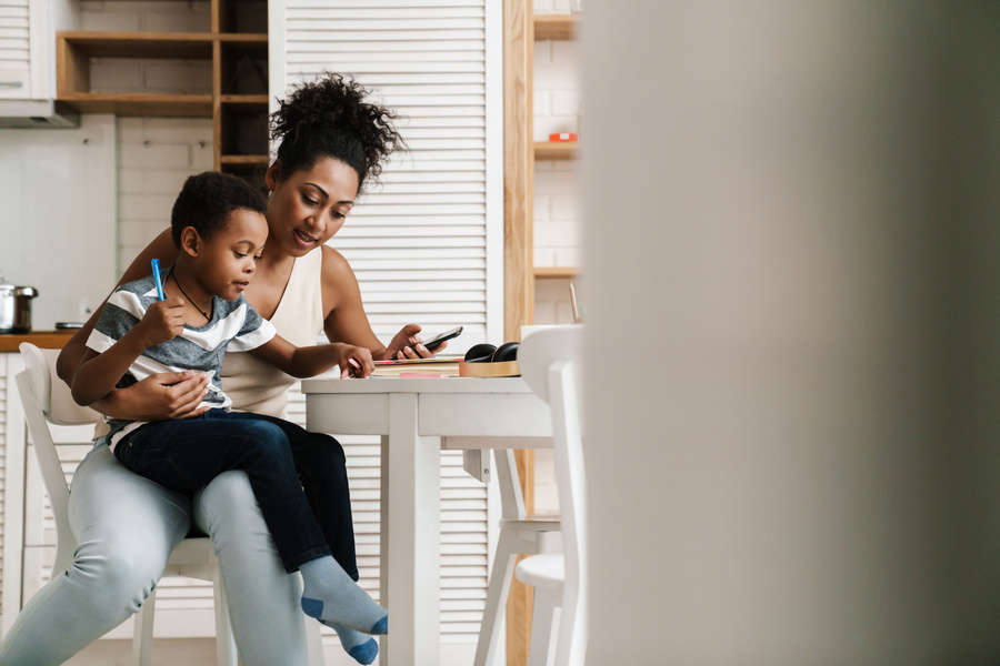 Black mother and son writing and using mobile phone while sitting together at home