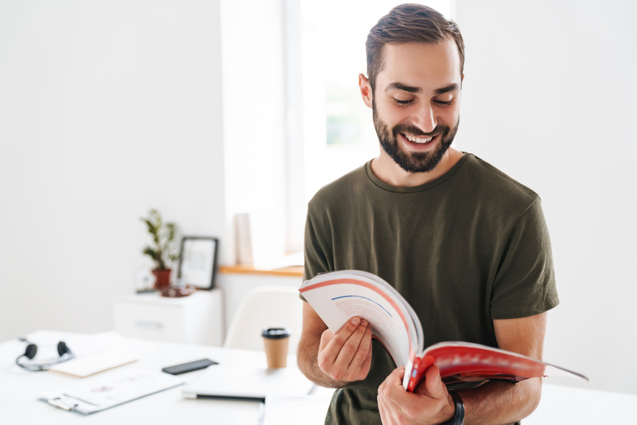 Image of caucasian happy man wearing casual t-shirt reading brochure and smiling while working in bright office