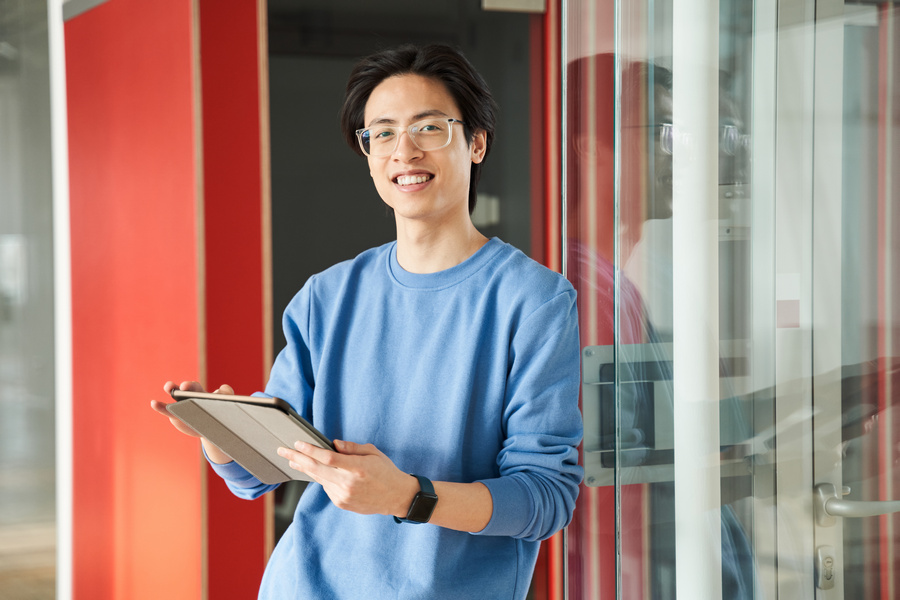Portrait of a happy smart male student wearing glasses using his tablet in university while standing at the hall near the red doors at the break. One person. Education during pandemic concept